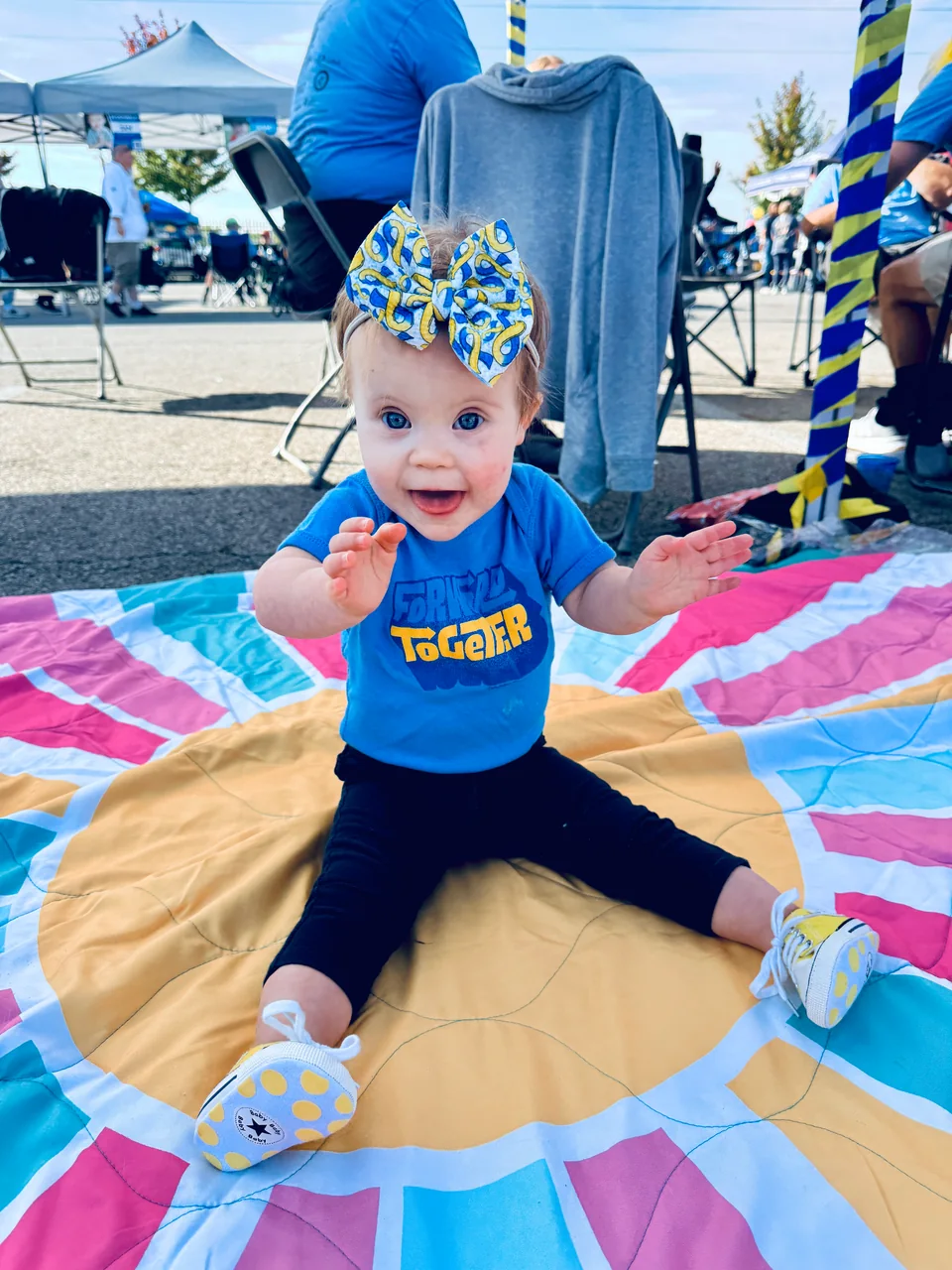 A smiling child in a blue shirt and a blue-and-yellow bow sits on a colorful quilt at an outdoor gathering, hands raised.